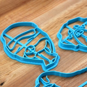 May include: A set of four blue plastic cookie cutters shaped like faces. The cutters are on a wooden surface.