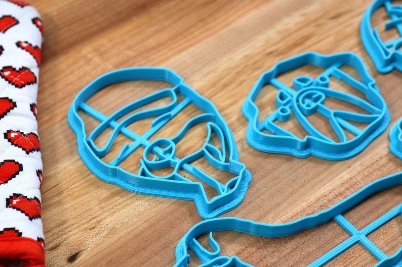 May include: A set of four blue plastic cookie cutters shaped like faces. The cutters are on a wooden surface.