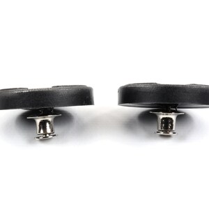 May include: Two black plastic buttons with a silver metal post and a small metal ring. The buttons are round and have a slightly raised center.