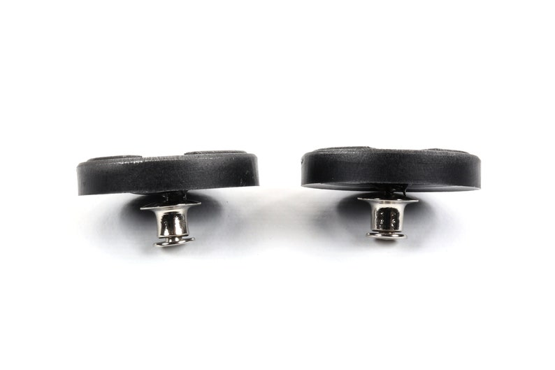 May include: Two black plastic buttons with a silver metal post and a small metal ring. The buttons are round and have a slightly raised center.
