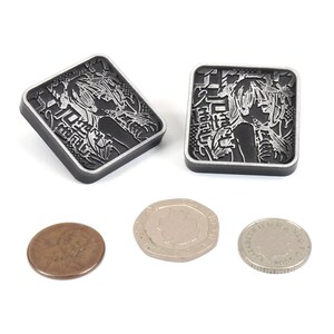 Puede incluir: Dos amuletos cuadrados de metal negro con una imagen plateada de una mujer con el pelo largo y texto japon&eacute;s. Los amuletos son aproximadamente del tama&ntilde;o de una moneda de 20 c&eacute;ntimos de euro.