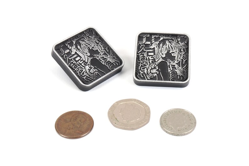 Puede incluir: Dos amuletos cuadrados de metal negro con una imagen plateada de una mujer con el pelo largo y texto japon&eacute;s. Los amuletos son aproximadamente del tama&ntilde;o de una moneda de 20 c&eacute;ntimos de euro.