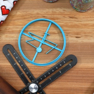 May include: A blue plastic cookie cutter shaped like a stylized airplane with a circle around it. The cutter is on a wooden surface with a ruler and a rolling pin.