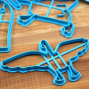 May include: A set of blue plastic cookie cutters in the shapes of a bird, a heart, and a cross. The cutters are on a wooden surface.
