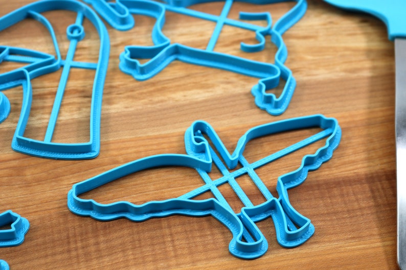 May include: A set of blue plastic cookie cutters in the shapes of a bird, a heart, and a cross. The cutters are on a wooden surface.