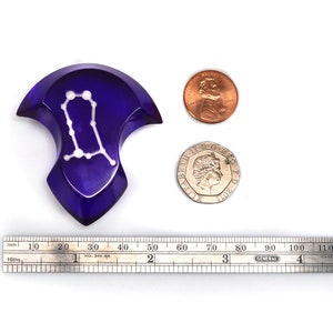 May include: A purple glass Gemini zodiac sign with white stars, measuring approximately 5 cm in diameter. The sign is next to a ruler and two coins for scale.