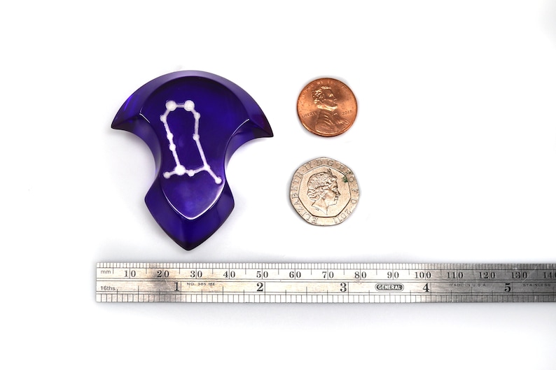 May include: A purple glass Gemini zodiac sign with white stars, measuring approximately 5 cm in diameter. The sign is next to a ruler and two coins for scale.
