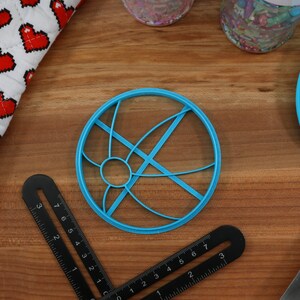 May include: A blue plastic cookie cutter shaped like a circle with a design of intersecting lines. The cutter is on a wooden surface.