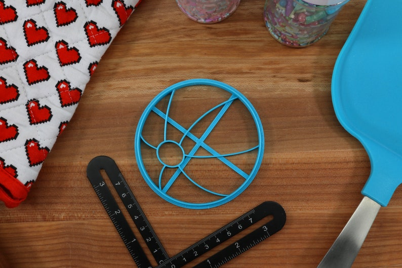 May include: A blue plastic cookie cutter shaped like a circle with a design of intersecting lines. The cutter is on a wooden surface.