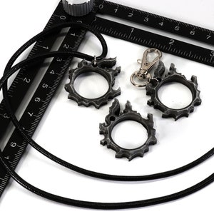 Puede incluir: Dos colgantes de metal de color plateado con un dise&ntilde;o de llama, uno en un collar de cord&oacute;n negro y el otro en un llavero plateado.
