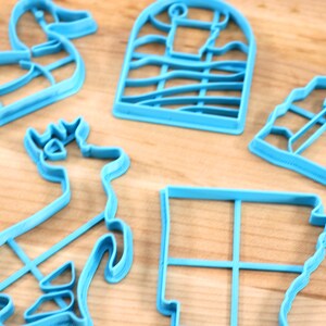 Op de afbeelding: Vijf blauwe plastic koekjesvormen in verschillende vormen: een eend, een hert, een huis, een gebouw en een vierkant.