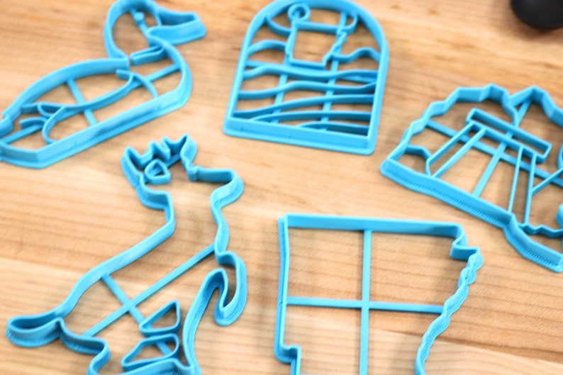 Op de afbeelding: Vijf blauwe plastic koekjesvormen in verschillende vormen: een eend, een hert, een huis, een gebouw en een vierkant.