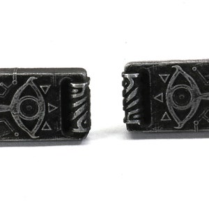 May include: A pair of silver-toned metal cufflinks featuring a detailed design of a stylized eye with a triangle pattern. The cufflinks are shaped like a small book.