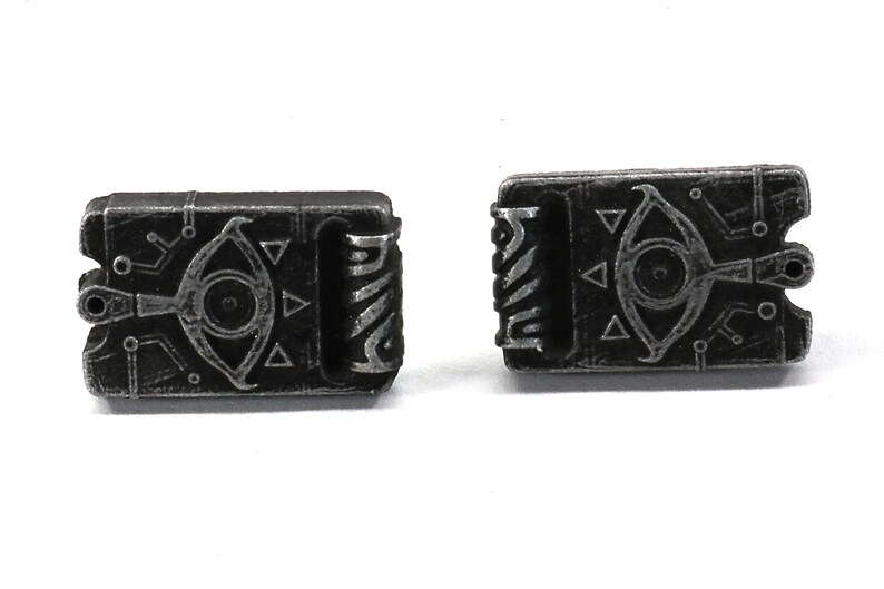 May include: A pair of silver-toned metal cufflinks featuring a detailed design of a stylized eye with a triangle pattern. The cufflinks are shaped like a small book.