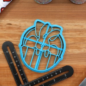 May include: A blue plastic cookie cutter shaped like a bunny rabbit inside a circle. The cutter is on a wooden surface.