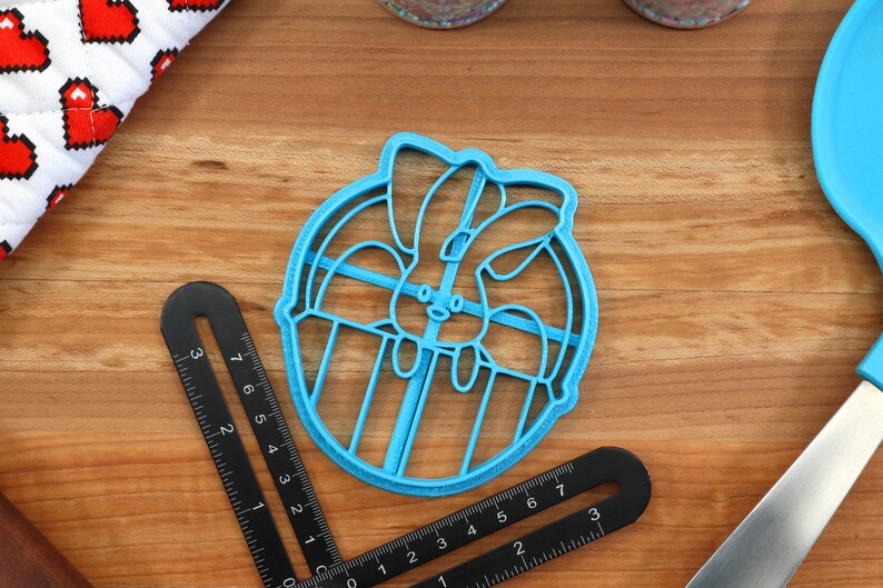 May include: A blue plastic cookie cutter shaped like a bunny rabbit inside a circle. The cutter is on a wooden surface.