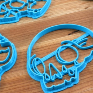 May include: Three blue plastic cookie cutters shaped like skulls. The cutters are on a wooden surface.
