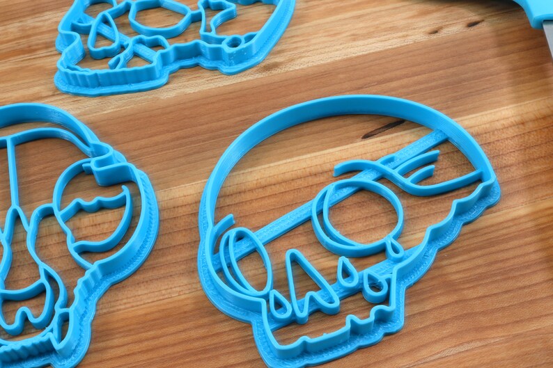 May include: Three blue plastic cookie cutters shaped like skulls. The cutters are on a wooden surface.