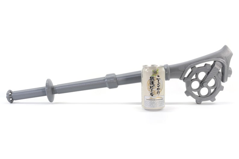 May include: A gray plastic can opener with a long handle and a gear-like mechanism. The opener is designed to open cans of beer. A can of beer is shown next to the opener for scale.