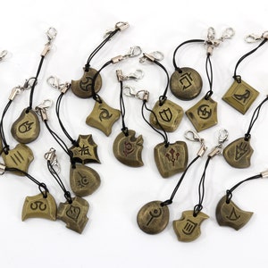 Może przedstawiać: Zestaw 17 złotych charm&oacute;w z r&oacute;żnymi symbolami i wzorami. Każdy charm jest przymocowany do czarnego sznurka ze srebrnym zapięciem.