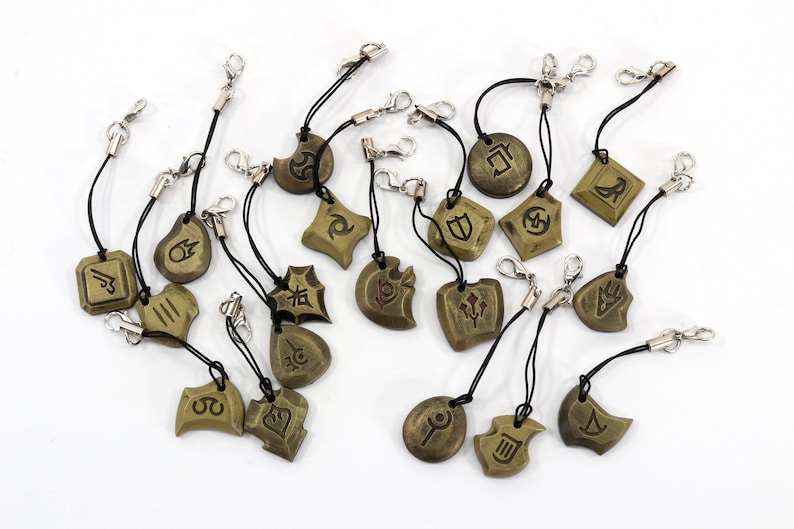 Może przedstawiać: Zestaw 17 złotych charm&oacute;w z r&oacute;żnymi symbolami i wzorami. Każdy charm jest przymocowany do czarnego sznurka ze srebrnym zapięciem.