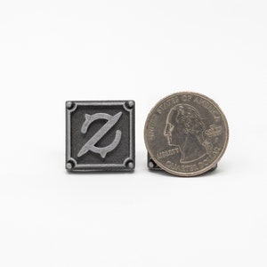 May include: A silver metal pin with a stylized letter Z in the center. The pin is about the size of a US quarter.