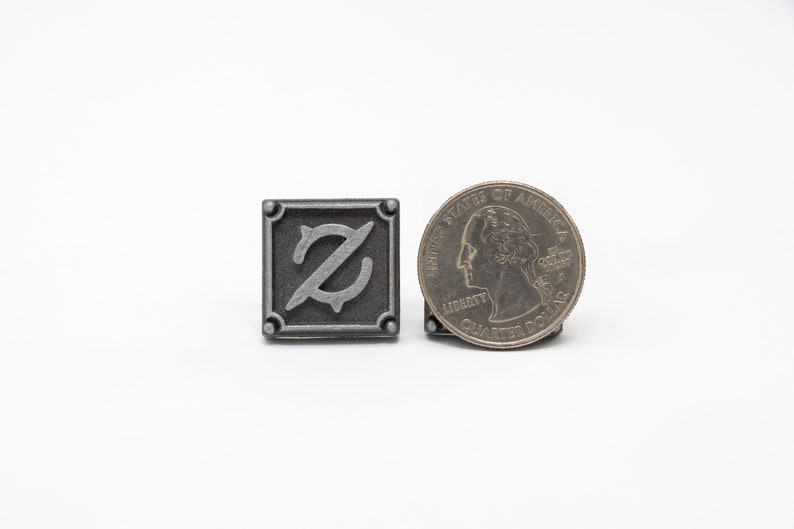 May include: A silver metal pin with a stylized letter Z in the center. The pin is about the size of a US quarter.