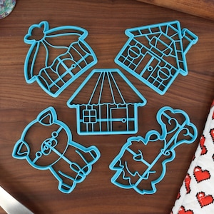 Puede incluir: Un conjunto de cinco cortadores de galletas de plástico azul con forma de casa, cerdo, árbol y dos estilos diferentes de casas. Los cortadores están sobre una superficie de madera.
