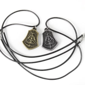 May include: Two necklaces with pendants featuring a stylized symbol. One pendant is gold-toned, the other is silver-toned. Both pendants are on black cords.