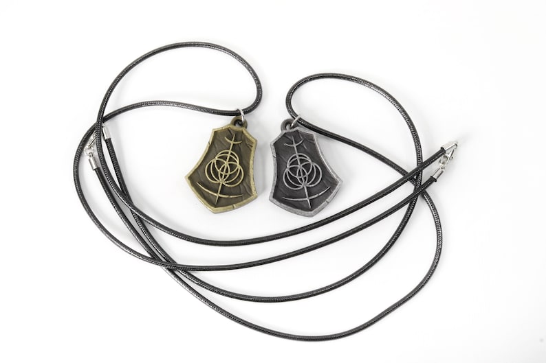 May include: Two necklaces with pendants featuring a stylized symbol. One pendant is gold-toned, the other is silver-toned. Both pendants are on black cords.