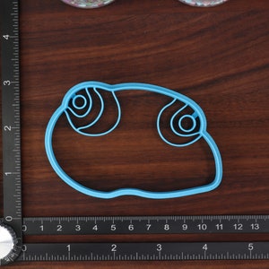 May include: A light blue plastic cookie cutter shaped like a cartoon eye with a double-circle design.