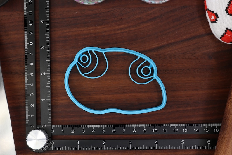 May include: A light blue plastic cookie cutter shaped like a cartoon eye with a double-circle design.