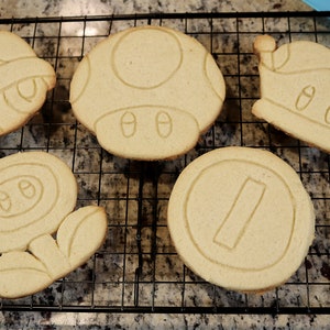 May include: Five different cookie cutters shaped like iconic video game characters. The cutters are made of metal and are in the shape of a mushroom, a turtle shell, a flower, a coin, and a crown.