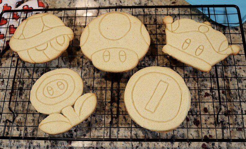 May include: Five different cookie cutters shaped like iconic video game characters. The cutters are made of metal and are in the shape of a mushroom, a turtle shell, a flower, a coin, and a crown.