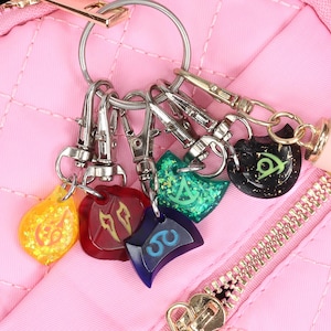 May include: A pink quilted bag with a zipper closure and a set of six colorful keychains attached to it. The keychains are made of metal and have different designs, including a yellow star, a red heart, a blue crescent moon, a green clover, a purple flower, and a black and green symbol.