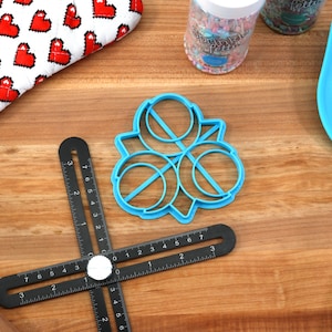 May include: Blue plastic cookie cutter shaped like a four-leaf clover with three circles in the center. The cutter is on a wooden surface with a black ruler, a rolling pin, and a blue spatula.
