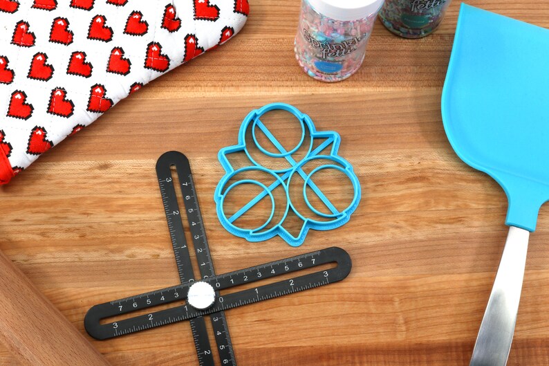 May include: Blue plastic cookie cutter shaped like a four-leaf clover with three circles in the center. The cutter is on a wooden surface with a black ruler, a rolling pin, and a blue spatula.
