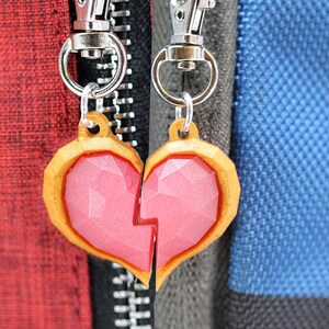 以下が含まれることがあります： 銀色のキーホルダーに付けられた、金色の縁取りのあるピンクのハート型のチャーム2つ。チャームは割れたハートのデザインです。