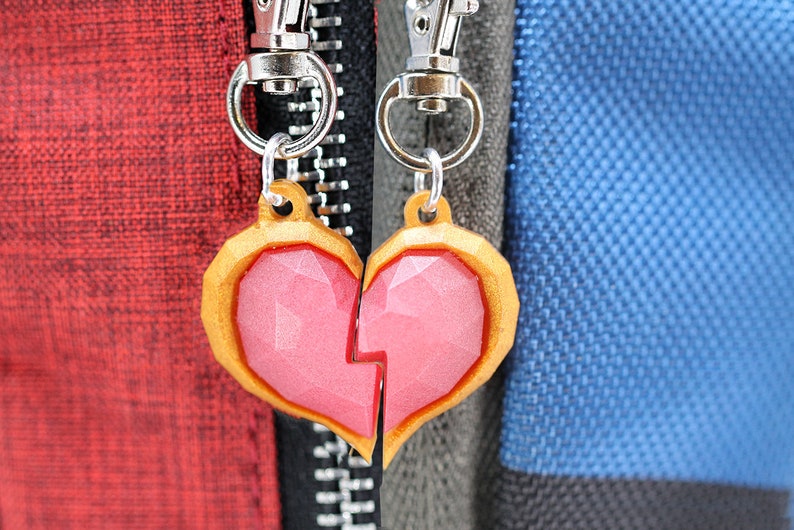 以下が含まれることがあります： 銀色のキーホルダーに付けられた、金色の縁取りのあるピンクのハート型のチャーム2つ。チャームは割れたハートのデザインです。