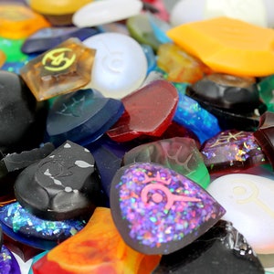 Puede incluir: Una colecci&oacute;n de p&uacute;as de guitarra &uacute;nicas y coloridas con varios dise&ntilde;os y texturas. Algunas p&uacute;as son transparentes, mientras que otras son opacas. Las p&uacute;as est&aacute;n hechas de diferentes materiales, incluyendo pl&aacute;stico, metal y madera.