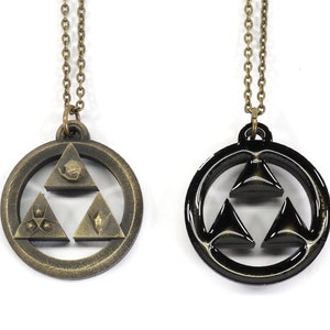 Puede incluir: Dos collares con un colgante de Trifuerza. Un collar tiene un colgante dorado con un dise&ntilde;o de Trifuerza detallado. El otro collar tiene un colgante negro con un dise&ntilde;o de Trifuerza simple.