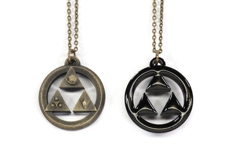 Puede incluir: Dos collares con un colgante de Trifuerza. Un collar tiene un colgante dorado con un dise&ntilde;o de Trifuerza detallado. El otro collar tiene un colgante negro con un dise&ntilde;o de Trifuerza simple.