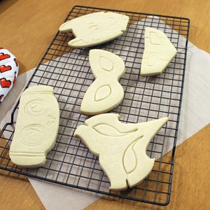 May include: A collection of freshly baked cookies in various shapes, including a jar, a bird, and abstract designs, resting on a black wire rack. A red and white oven mitt with pixelated hearts is visible on the left.