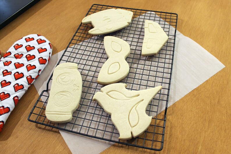 May include: A collection of freshly baked cookies in various shapes, including a jar, a bird, and abstract designs, resting on a black wire rack. A red and white oven mitt with pixelated hearts is visible on the left.
