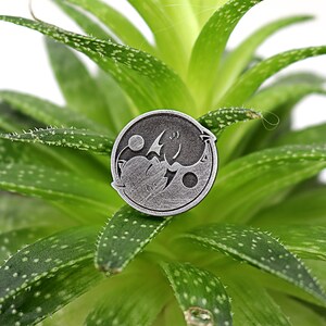 May include: Silver-toned yin yang pin with a dragon and a phoenix design. The pin is sitting on a green succulent plant.