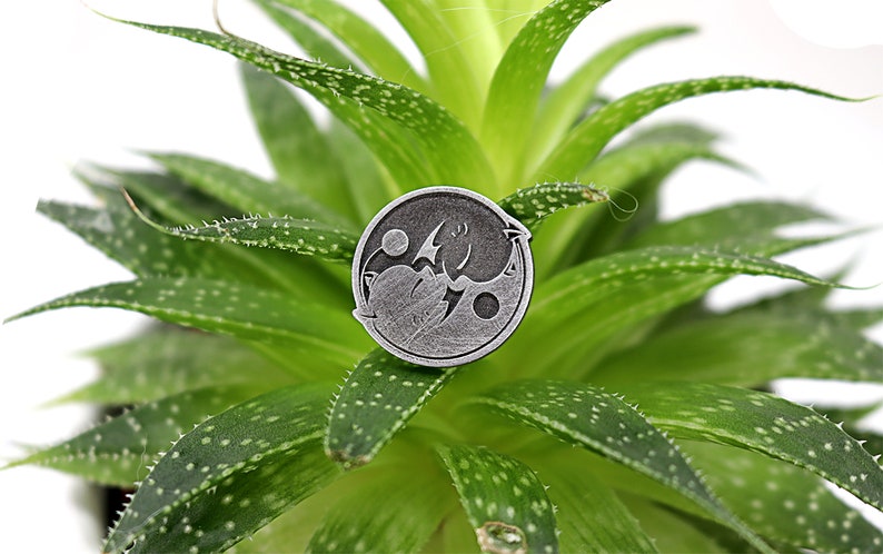 May include: Silver-toned yin yang pin with a dragon and a phoenix design. The pin is sitting on a green succulent plant.