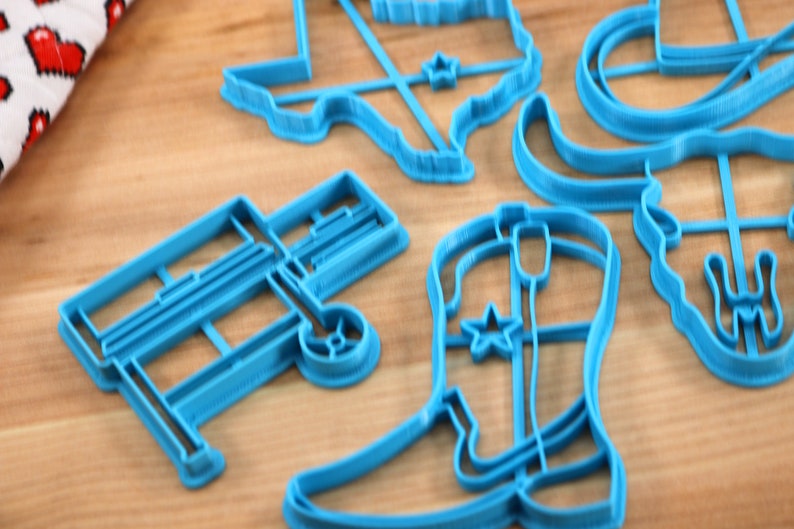 May include: A set of blue plastic cookie cutters shaped like a cowboy boot, a wagon, a longhorn steer, a cowboy hat, and the state of Texas.