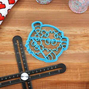May include: A blue plastic cookie cutter shaped like a cartoon pufferfish with a candy cane and stars. The cutter is on a wooden surface with a black ruler.