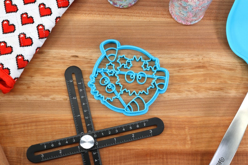 May include: A blue plastic cookie cutter shaped like a cartoon pufferfish with a candy cane and stars. The cutter is on a wooden surface with a black ruler.