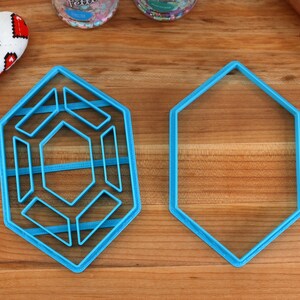 Op de afbeelding: Twee blauwe plastic koekjesvormen op een houten oppervlak. E&eacute;n vorm is een zeshoek, de andere is een complexere geometrische vorm met meerdere lagen.